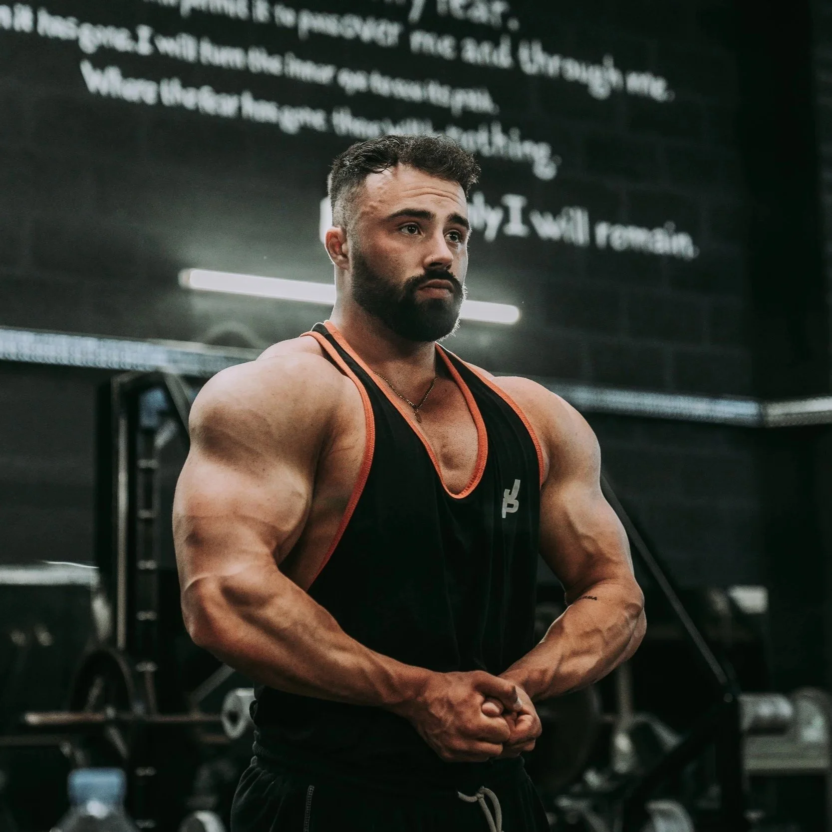 A muscular man with a beard and short hair posing in a gym, wearing a black and orange sleeveless shirt, with gym equipment and a black wall with writing in the background.