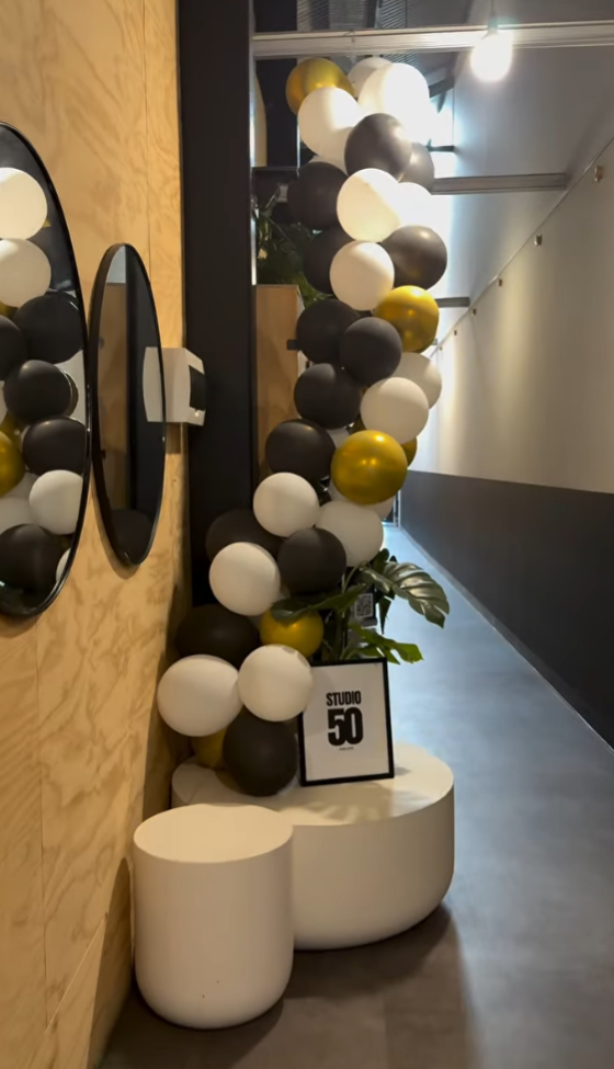 Decorative arrangement of black, white, and gold balloons with a small sign that reads 'STUDIO 50,' placed on a white curved stand inside a modern hallway.