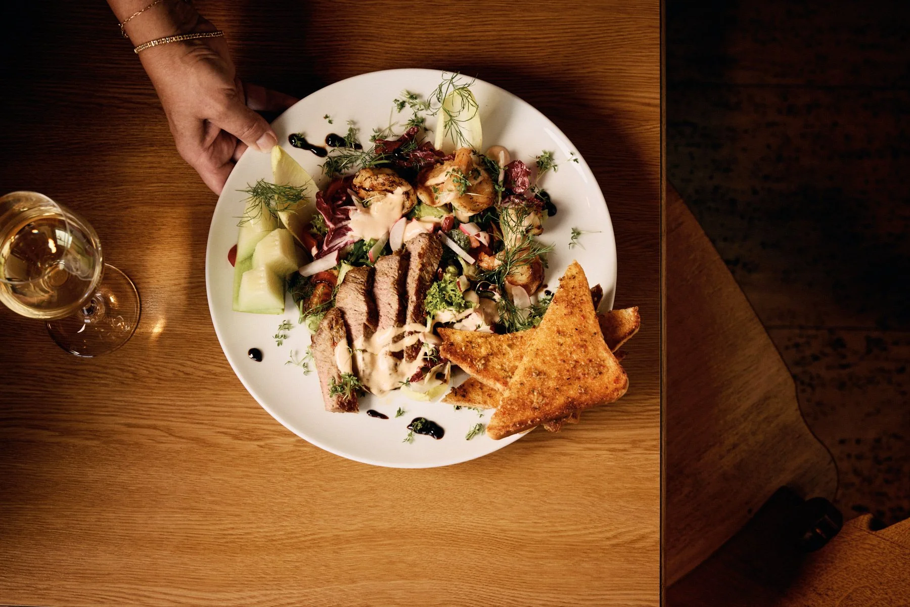 Auf einem Holzplatz steht ein großer Teller mit Salat, gebratenem Fleisch, gebratenen Toastscheiben und Melonenscheiben, neben einem Glas Weißwein.
