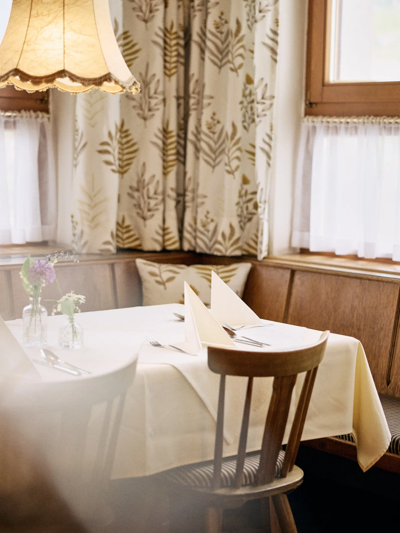 Ein festlich gedeckter Tisch in einem gemütlichen, traditionell eingerichteten Raum mit Holzvertäfelung, weißen Tischtüchern, Stoffservietten und kleinen Blumenvasen. Im Hintergrund sind Fenster mit Gardinen und eine Wand mit blumigem Muster.