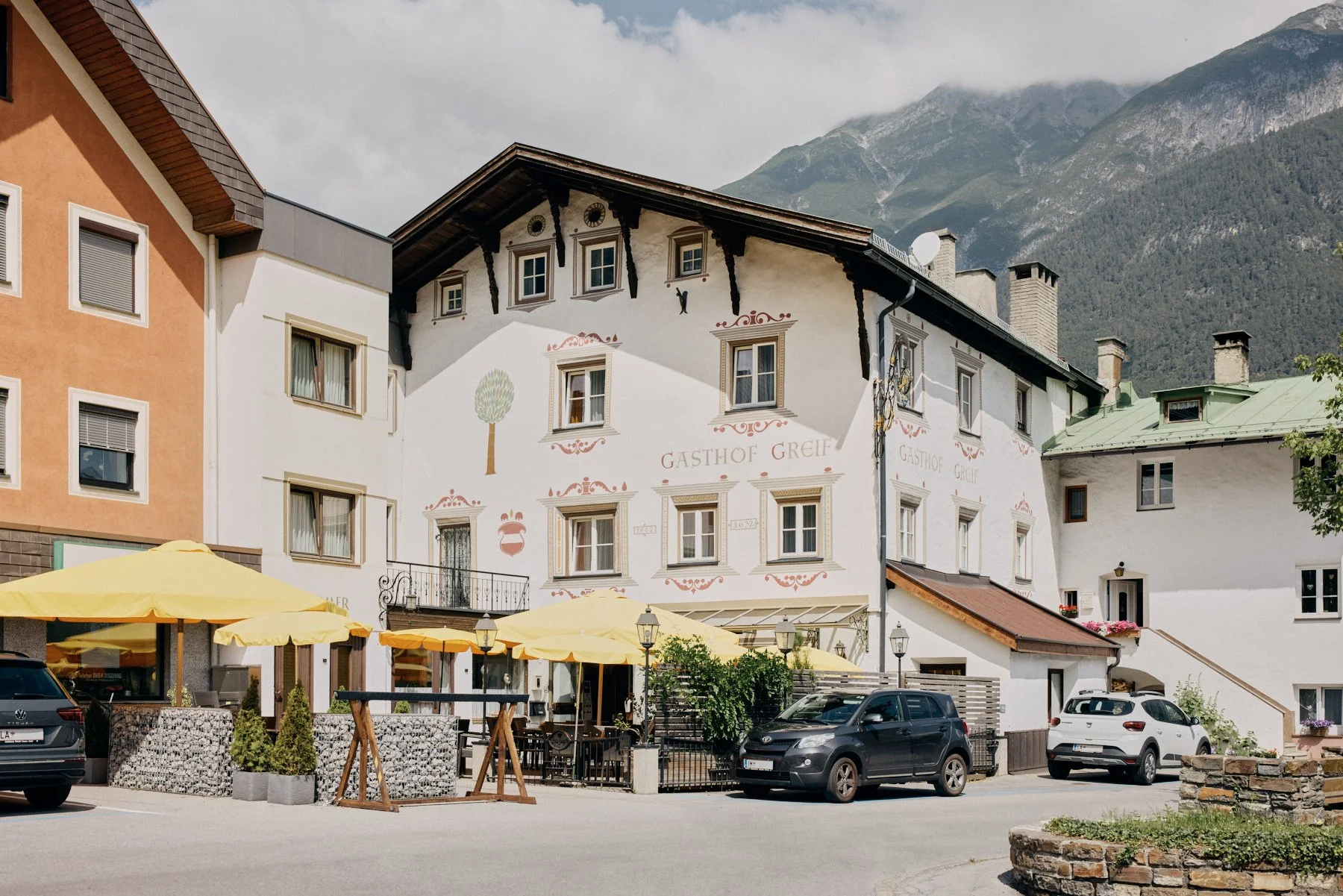 UnserTraditionelles Hotel Gasthof Greif in einer Landeck, Außenbereich mit Tischen und Sonnenschirmen, bewaldete Berge im Hintergrund.