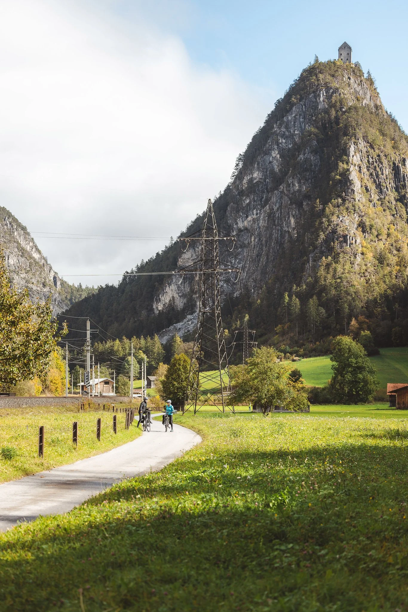 Eine grüne Wiese mit einem schlanken, gewundenen Weg, auf dem drei Radfahrer fahren. Im Hintergrund befinden sich hohe, gebrochene Felsen und bewaldete Hügel. Über der Wiese sind elektrische Leitungen und ein Sendemast sichtbar.