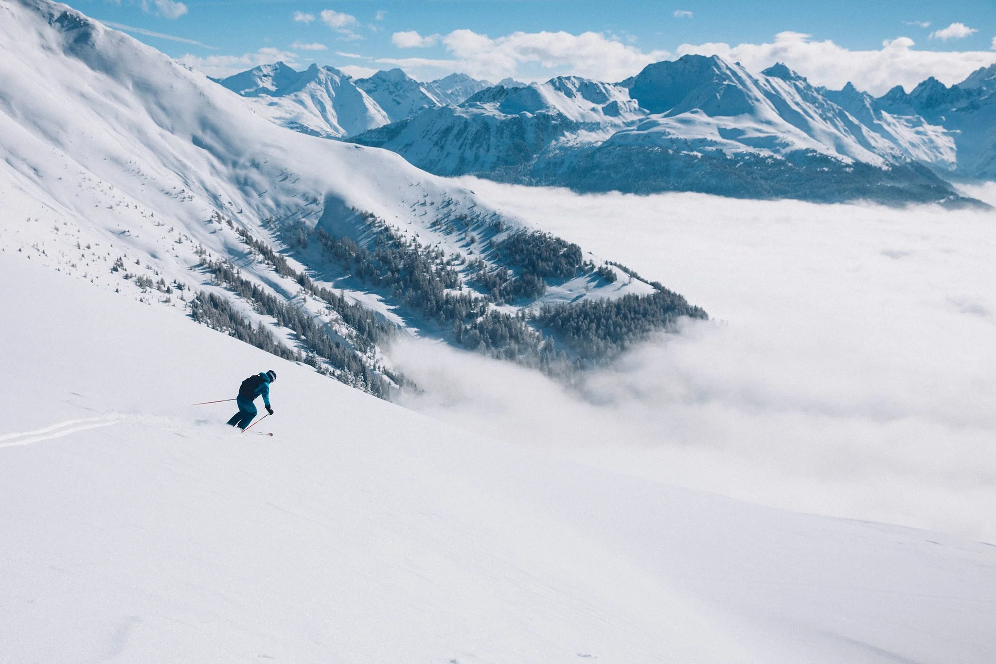 Ein Skifahrer in blauer Kleidung fährt durch tiefen Schnee in den Bergen, umgeben von schneebedeckten Gipfeln und Wolken in tiefer Talregion.