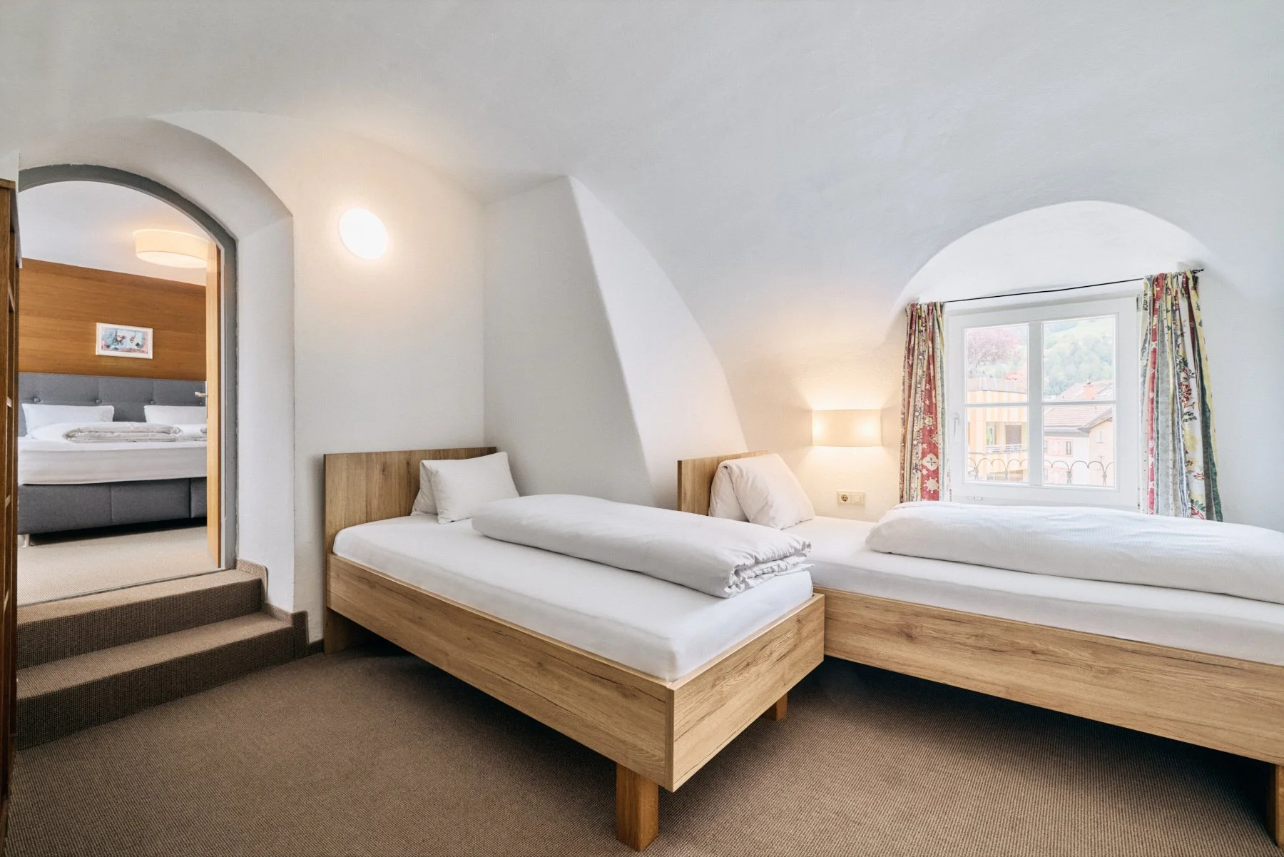 Schlafzimmer mit zwei Betten, einer Fensterfront mit bunten Vorhängen, weiße Wände und Decken, Holzmöbel, gemusterte Vorhänge, ein Durchgang zu einem anderen Schlafzimmer