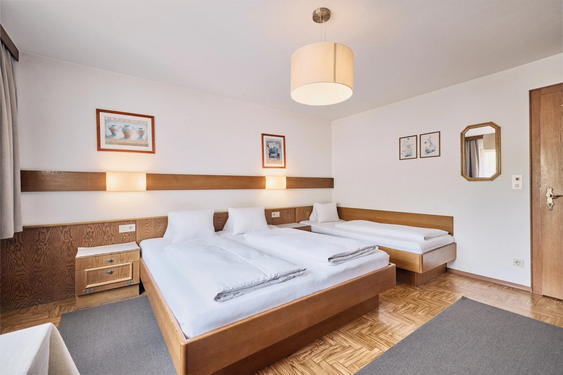 Ein Hotelzimmer mit drei Betten, Holzfußboden, Wandbildern und einem Deckenlampenschirm.