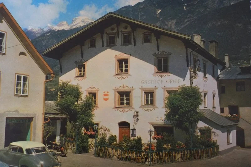 Ein malerisches Gasthaus namens 'Gasthof Greif' in einer Bergregion, umgeben von grünen Bäumen und Bergen im Hintergrund.