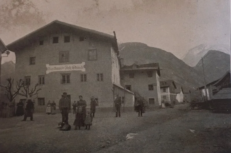 Schwarz-weiß-Foto eines Dorfes in den Bergen mit mehreren Gebäuden und einigen Menschen im Vordergrund.