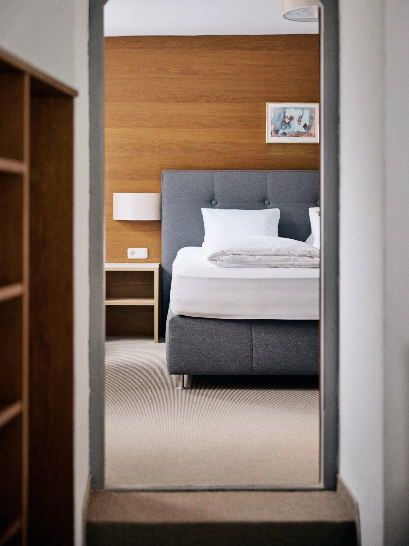 Blick in ein Hotelzimmer mit grauem Bett, weißen Kissen, Holz-Wand und einem kleinen Nachttisch
