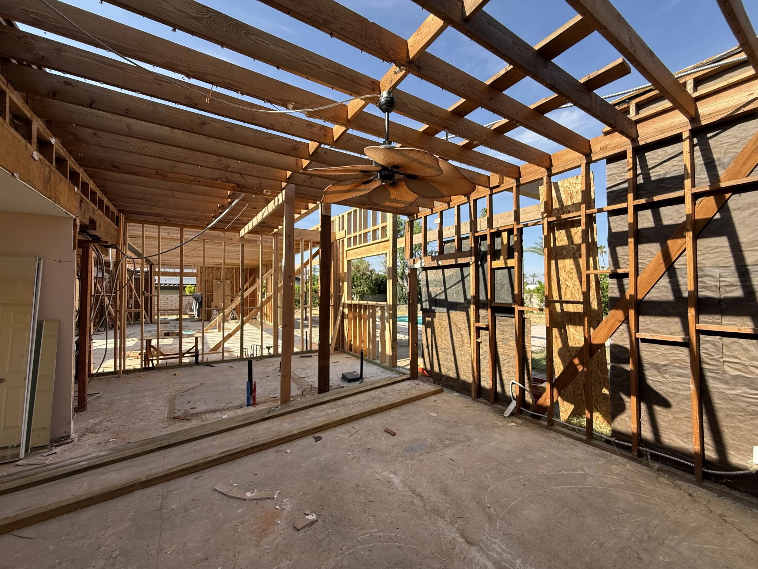Interior of a house under construction with exposed wooden framing, a ceiling fan, and a partially open wall showing an outdoor area.