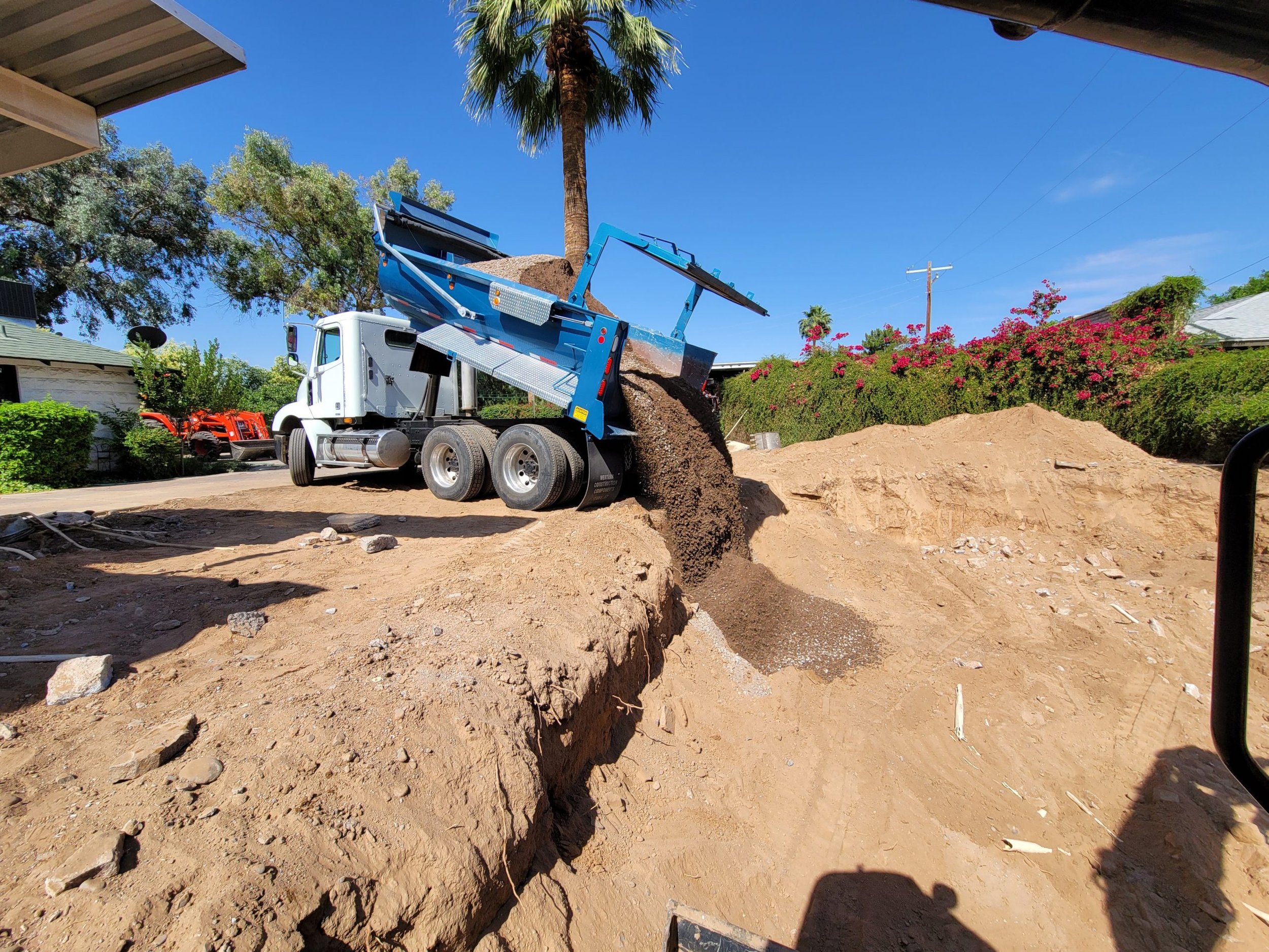 A white dump truck unloading dirt onto a construction site with dirt piles and trees in the background under a blue sky.