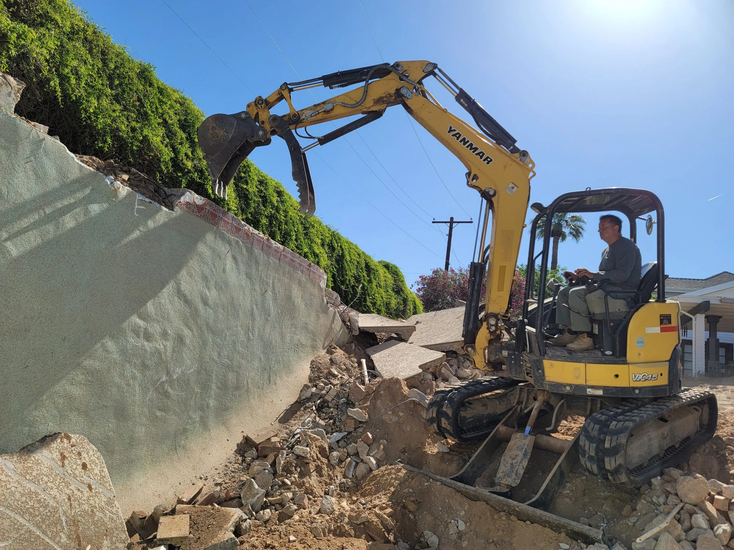 A man operating a small yellow excavator demolishing a concrete wall with green bushes on top.
