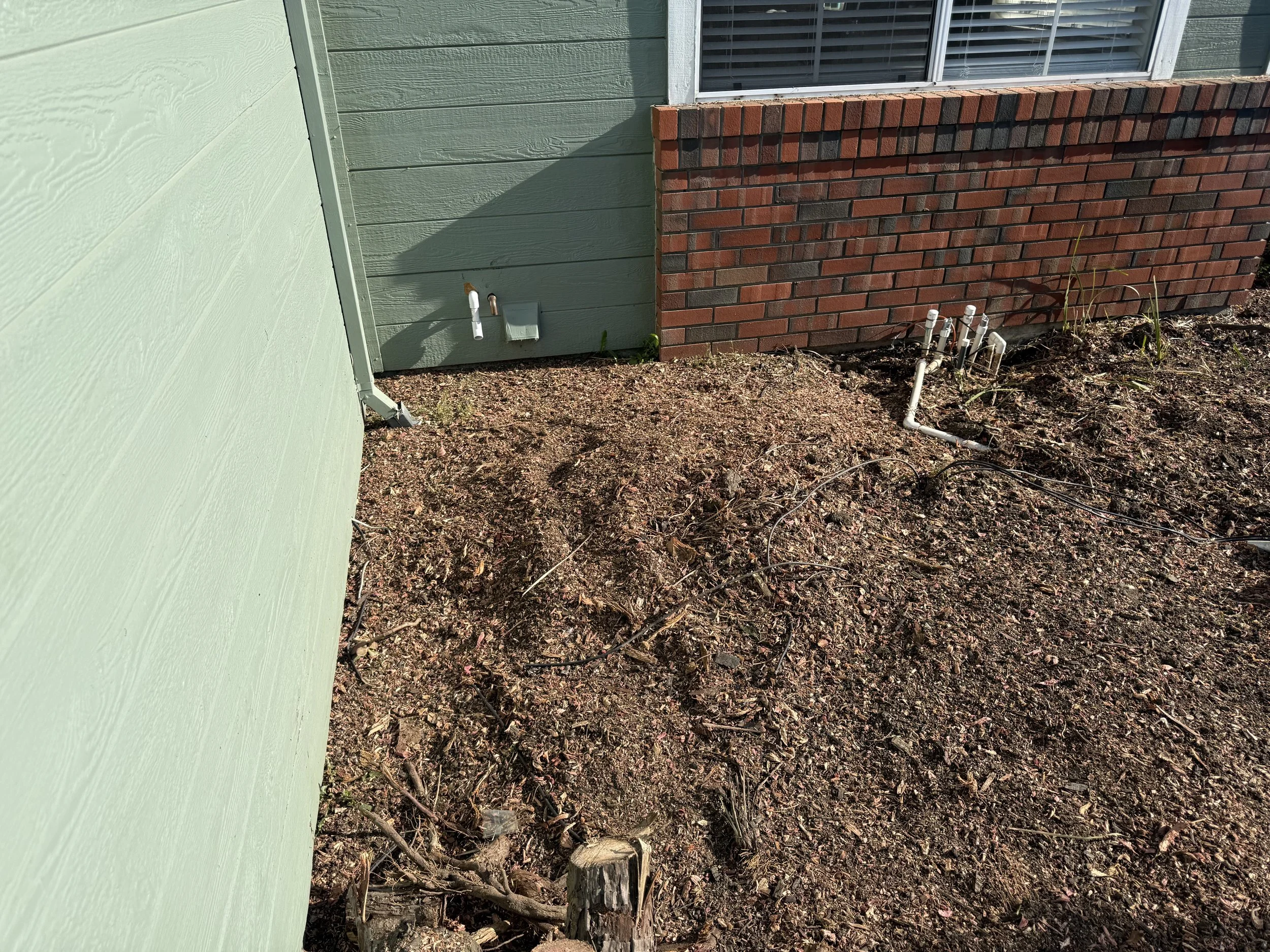 A patch of bare soil next to a house with green siding and a brick foundation, showing exposed water pipes, some small plants, and a few branches on the ground.