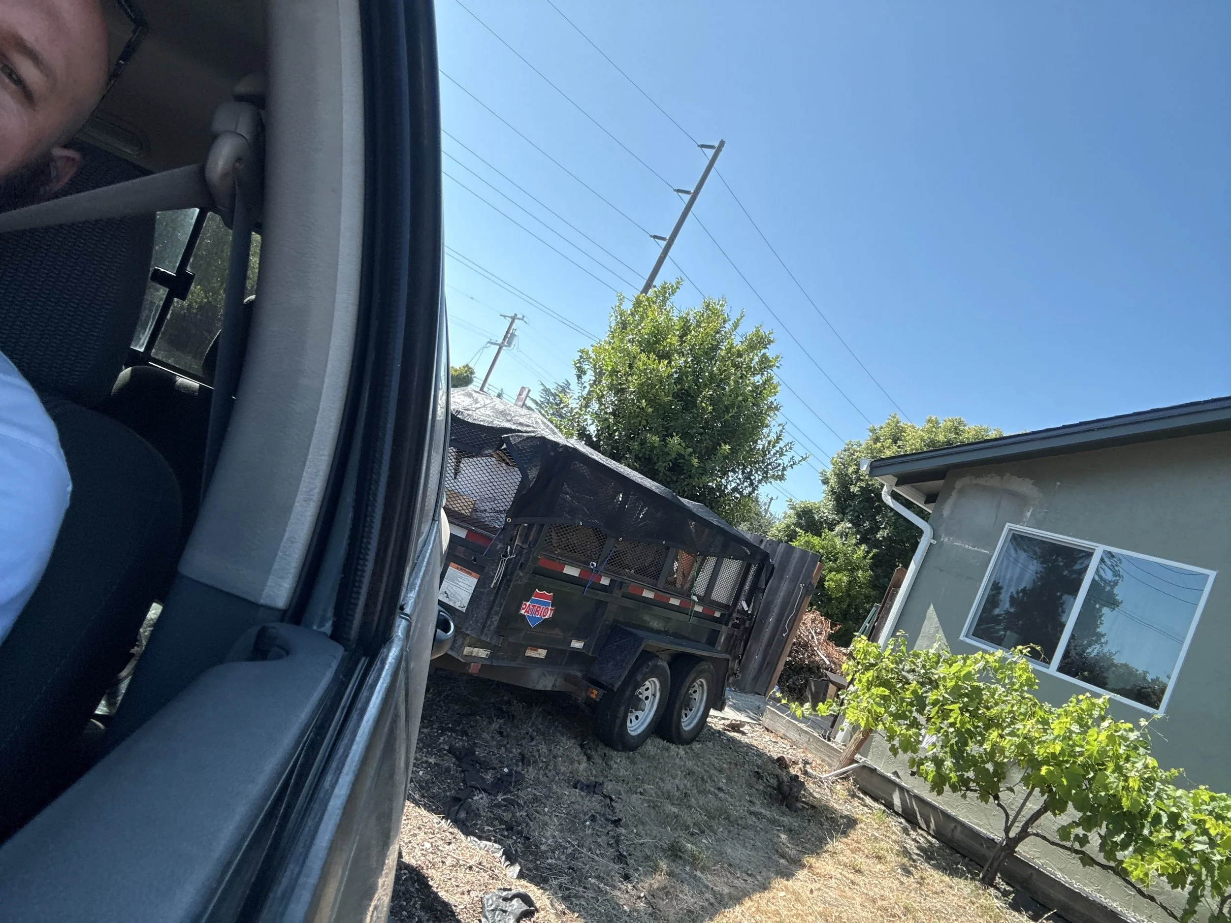 View from inside a vehicle showing a black trailer parked near a house with a green exterior, a large window, and some greenery in the yard under a clear blue sky.