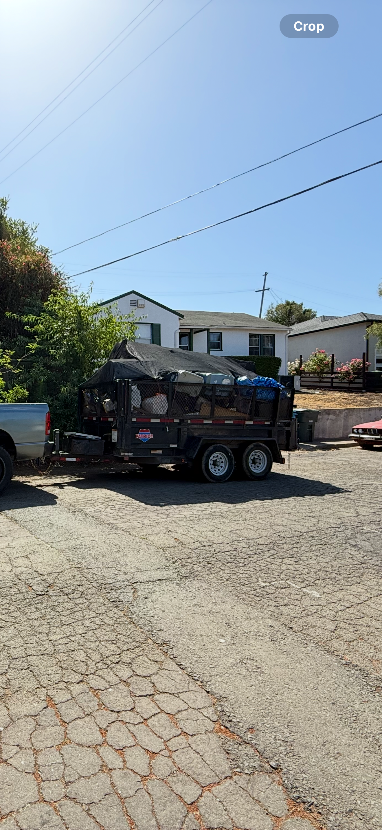 A black trailer filled with various items, covered with a tarp, is parked on a cracked asphalt driveway in front of residential houses.