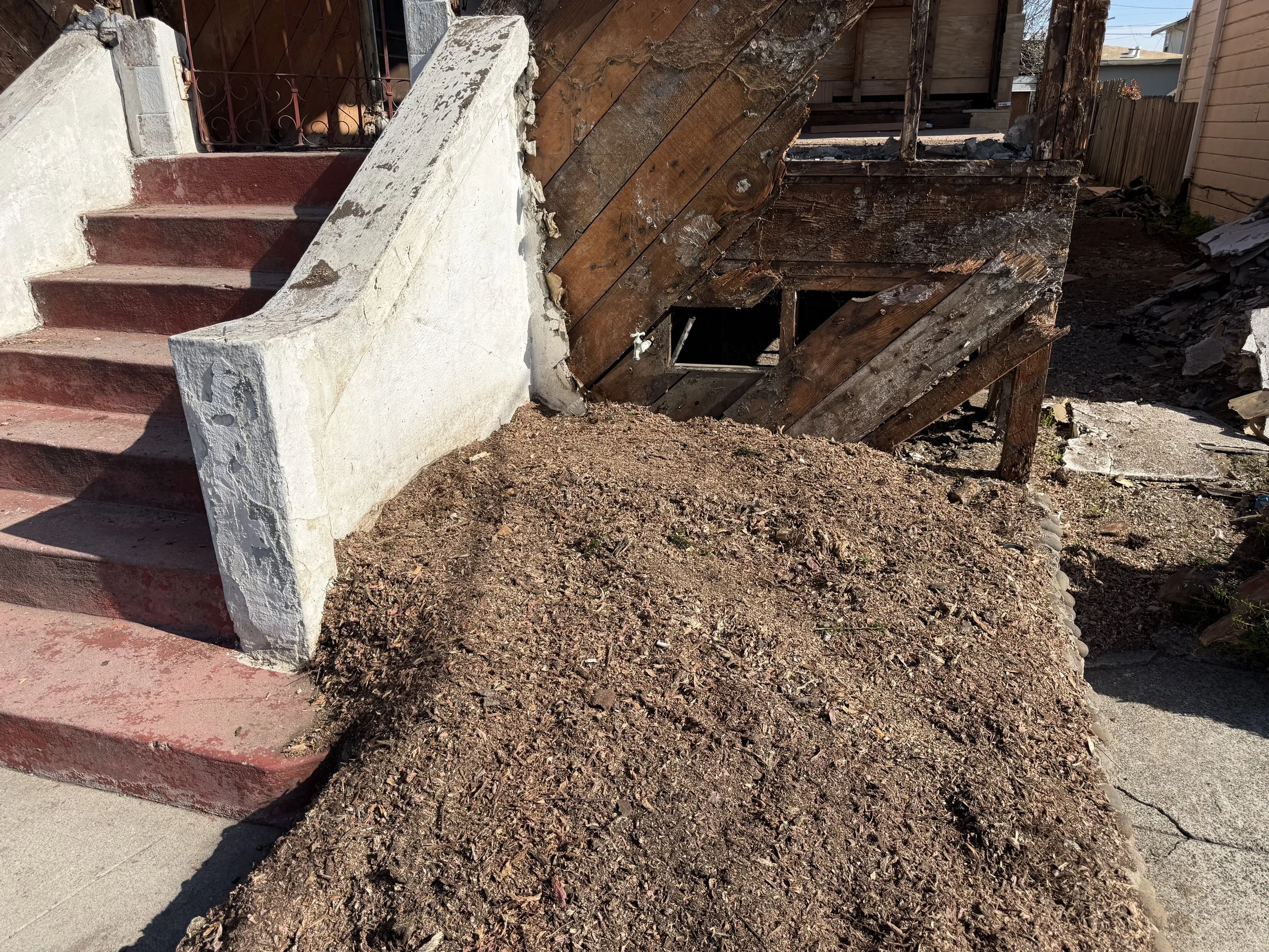 The image shows a house's exterior steps with some concrete and a mud pile at the base of the steps, along with a damaged wooden wall with exposed boards and a small hole.