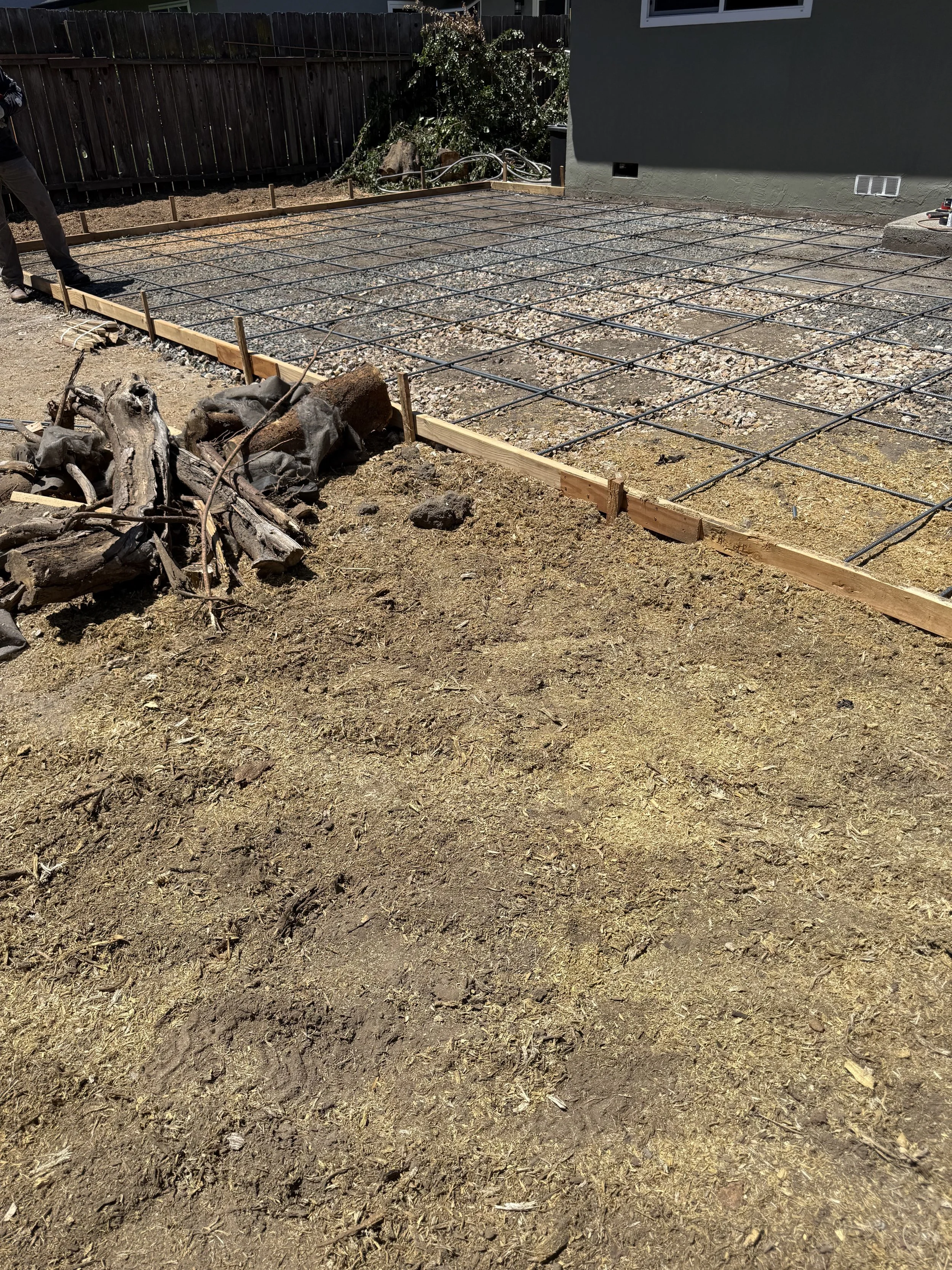 A backyard with a new concrete slab foundation in progress, reinforced with metal rebar, surrounded by wooden framing. There is a pile of logs and branches in the foreground and a dark wooden fence in the background.