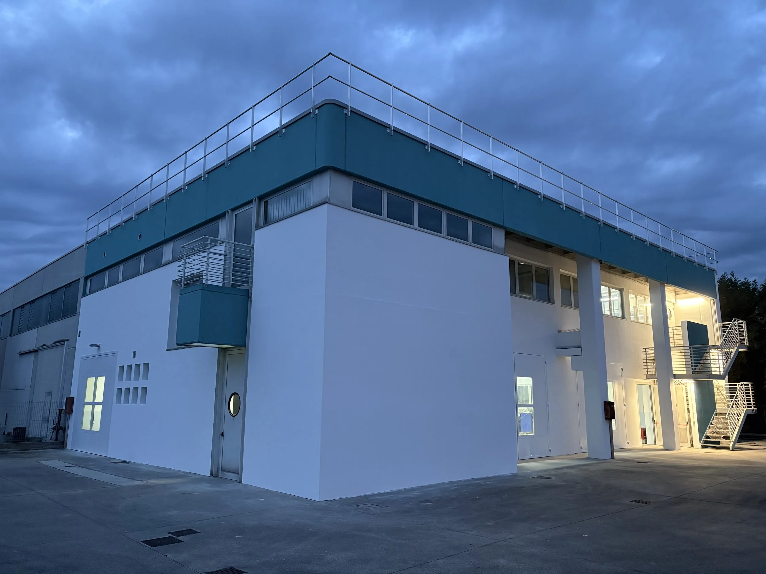 Edificio industrial con bianco e blu, luci accese, mure esterno e scale metalliche, al crepuscolo o sera.