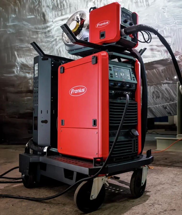 Apparecchiatura di saldatura Fronius con display digitale, montata su carrello con ruote, in un ambiente industriale.