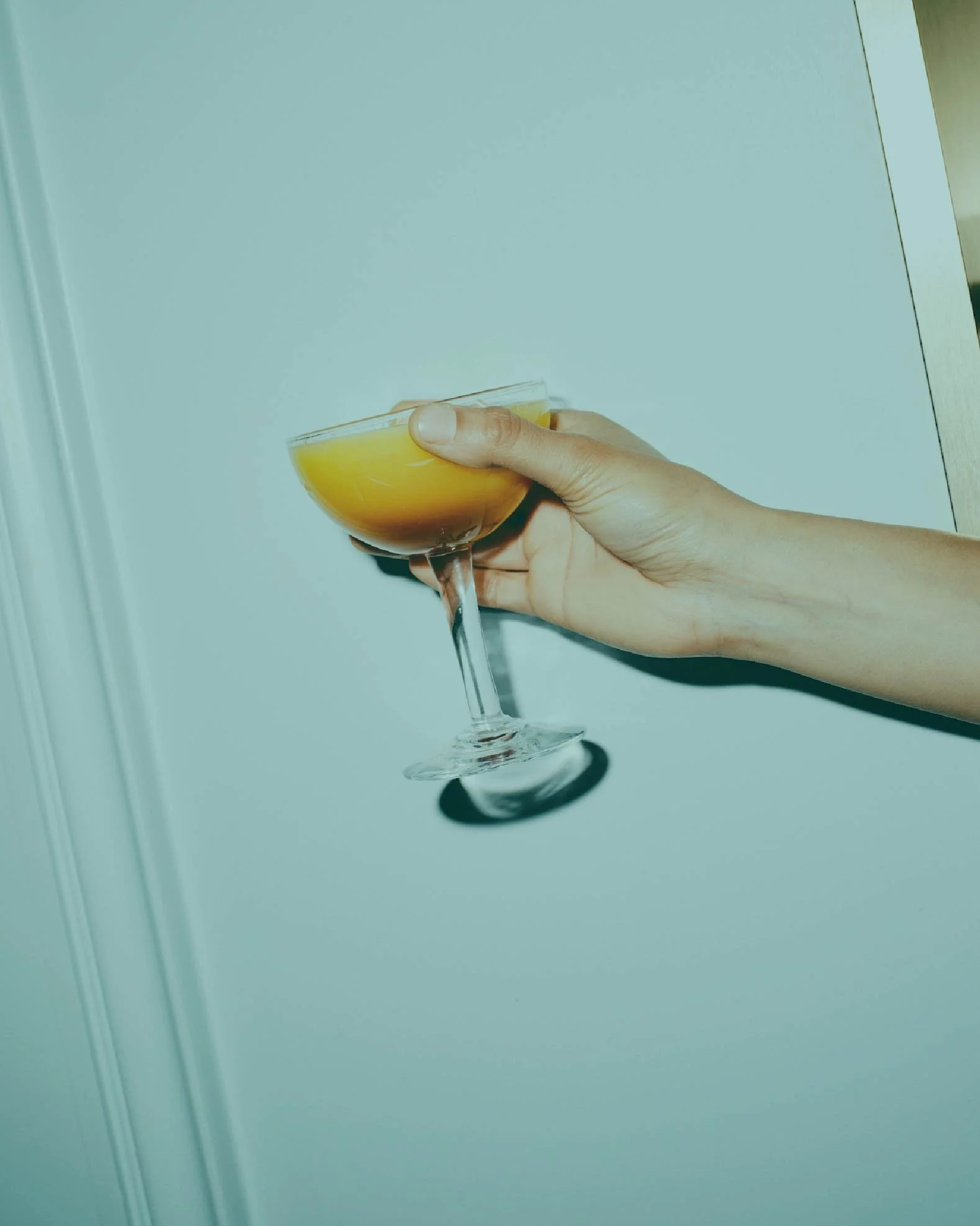 A hand holding a cocktail glass with a yellow-orange drink against a plain background.