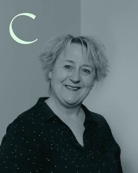 A smiling woman with short, curly hair wearing a black shirt with white polka dots, standing against a light-colored wall with a crescent moon symbol in the top left corner.