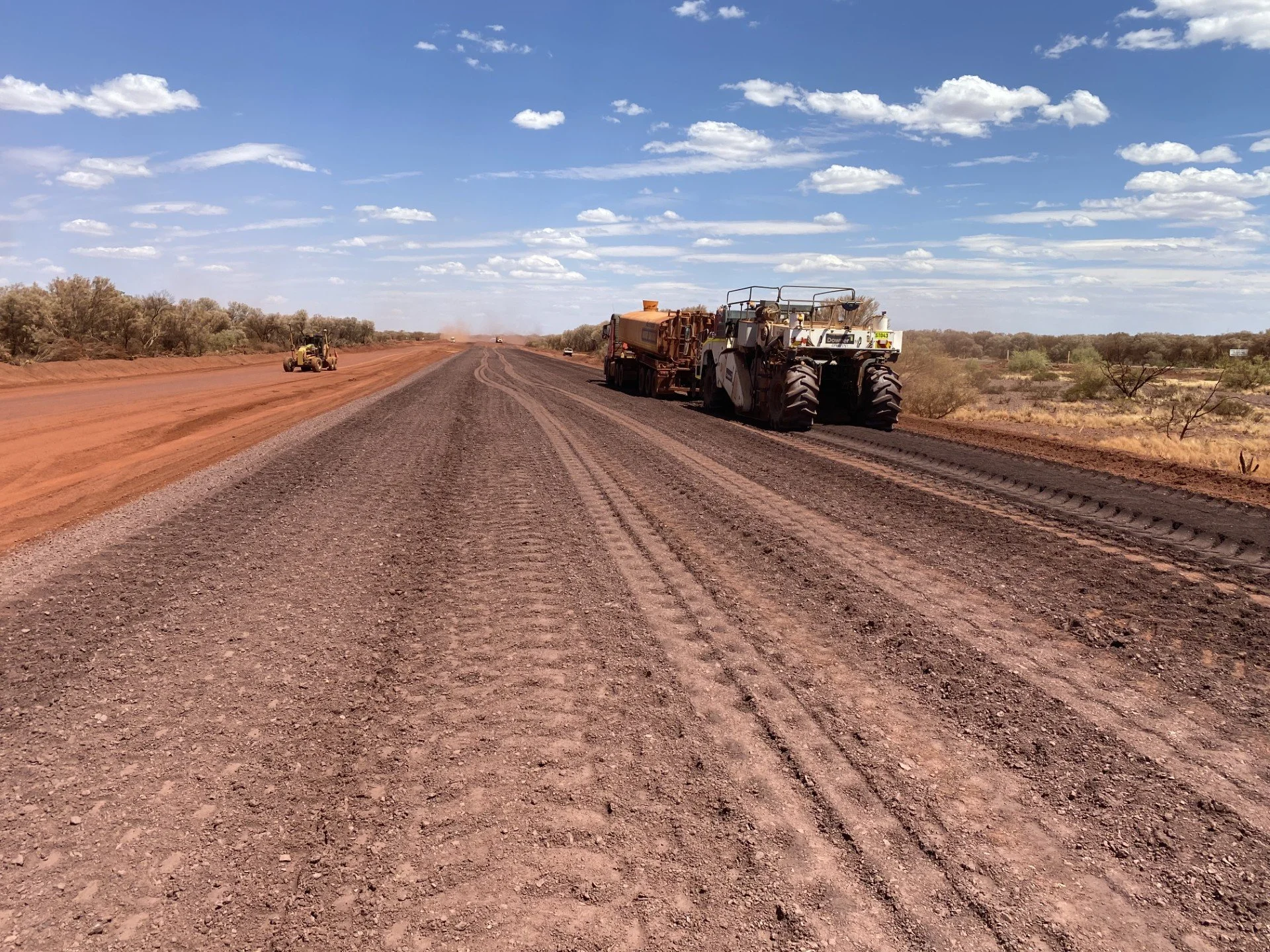 Construction equipment working on a dirt and gravel road in a desert landscape with dry shrubs, under a blue sky with scattered clouds.