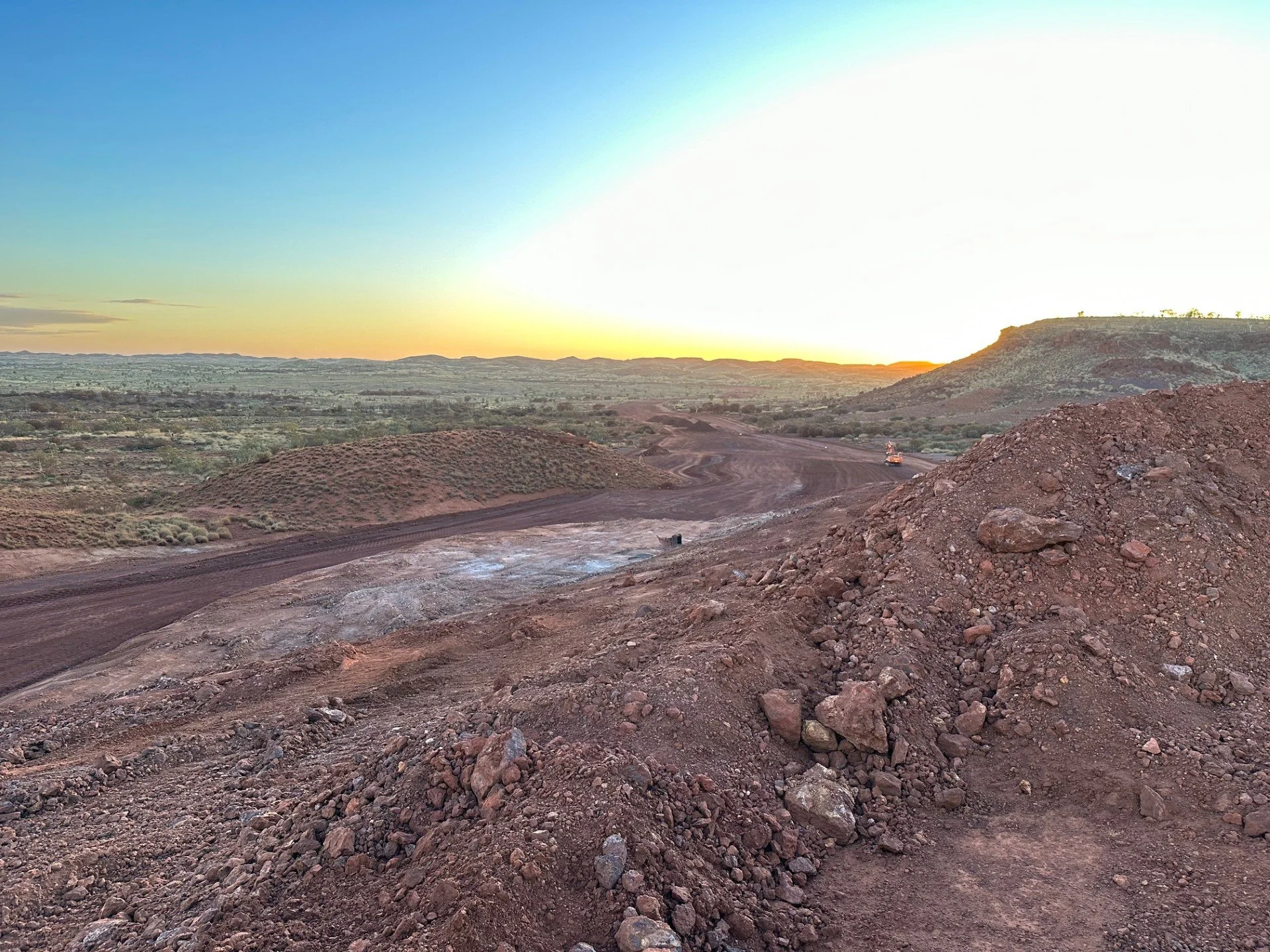 A construction site in a desert landscape at sunset, with dirt mounds, loose soil, and construction machinery in the distance.