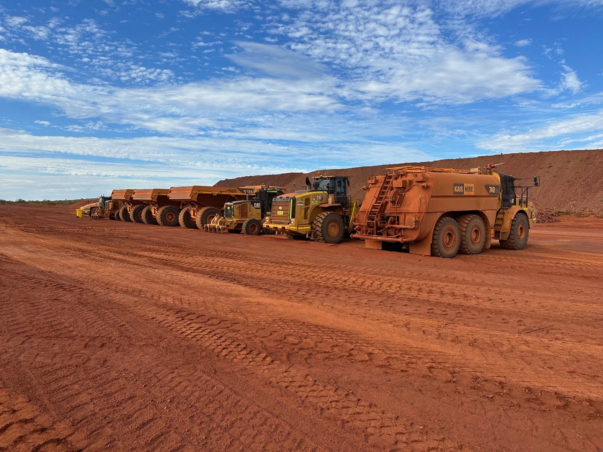 A row of large earth-moving dump trucks parked on red dirt under a blue sky with scattered clouds.