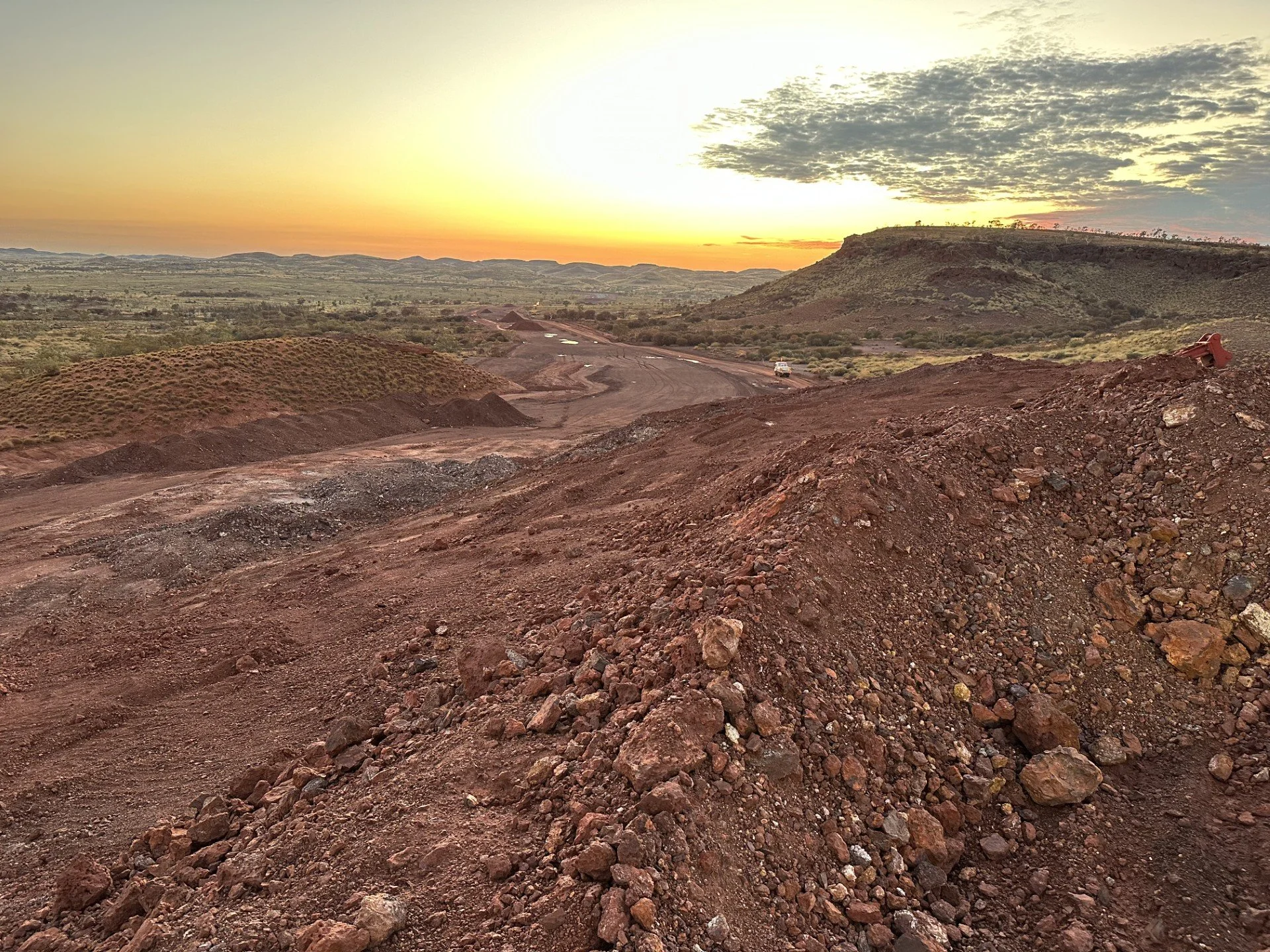 A landscape photo of a dirt road under construction in a semi-arid area with hills and sparse vegetation at sunset.