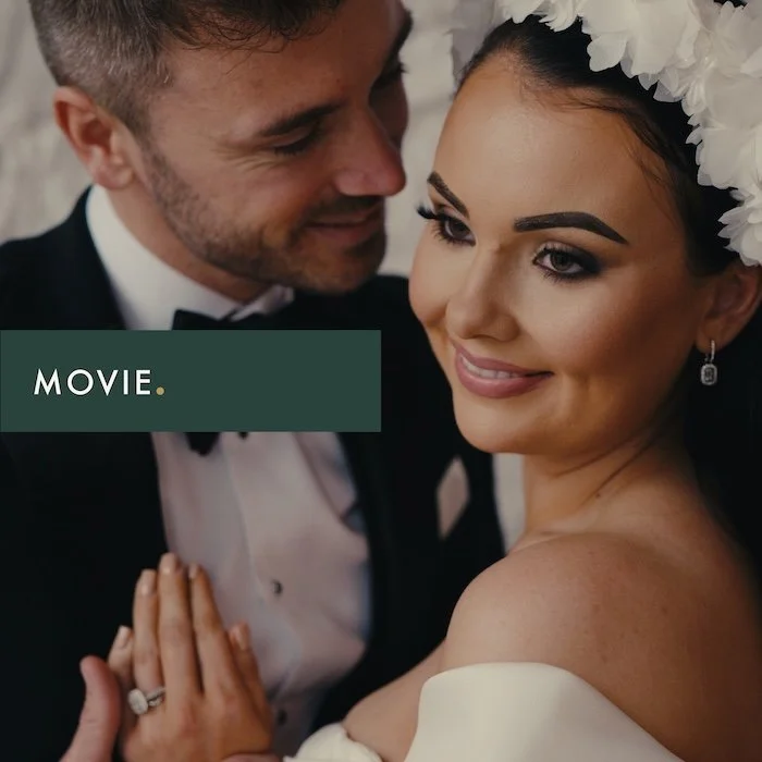 A wedding couple, a man and a woman, close together with the woman smiling, wearing a floral headpiece and earrings, the man in a tuxedo, in a romantic pose.