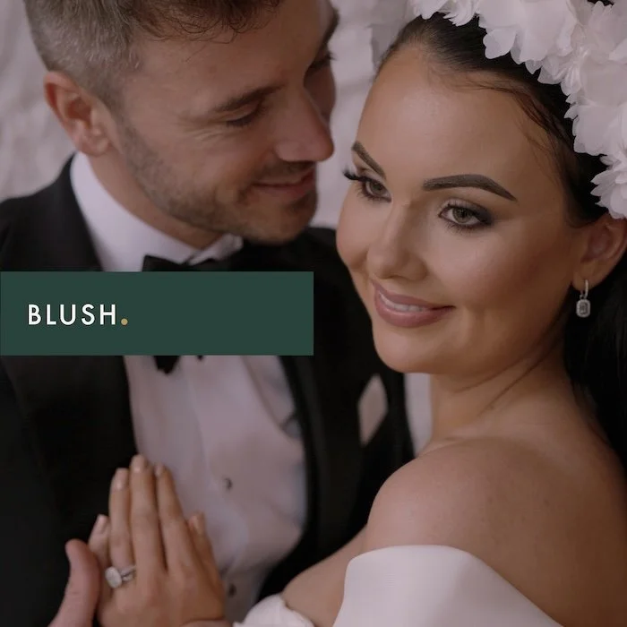 A bride and groom close together, smiling, with the bride wearing a white floral headpiece and earrings, and the groom in a tuxedo. The word 'BLUSH.' is written across the image.