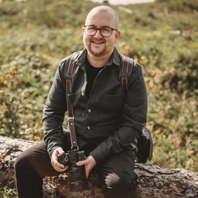 A man with glasses and a beard, wearing a black jacket, sitting on a log in a park or forested area, holding a camera with a strap around his neck, smiling at the camera.