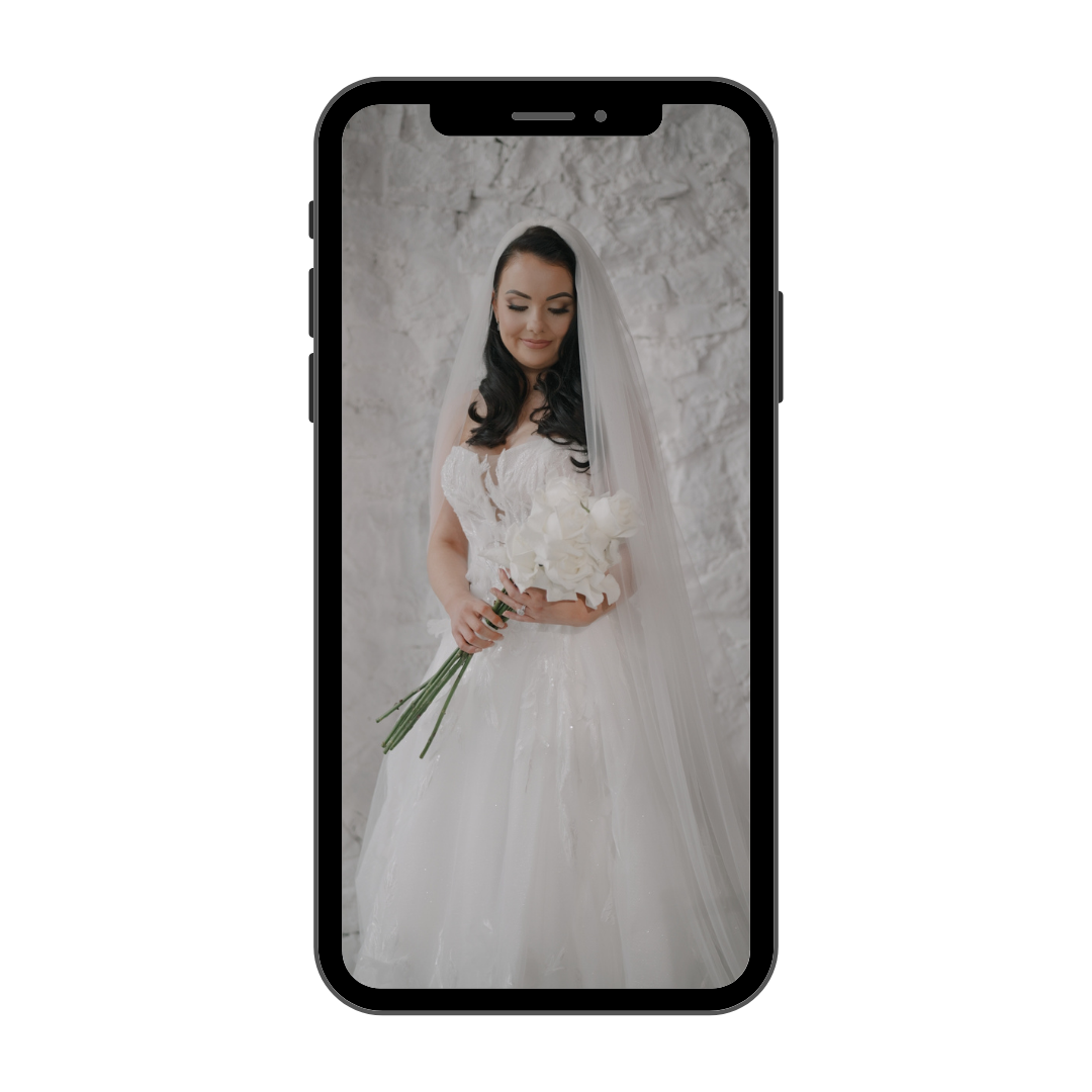 A woman in a white wedding dress holding a bouquet of white flowers, standing against a textured stone wall.