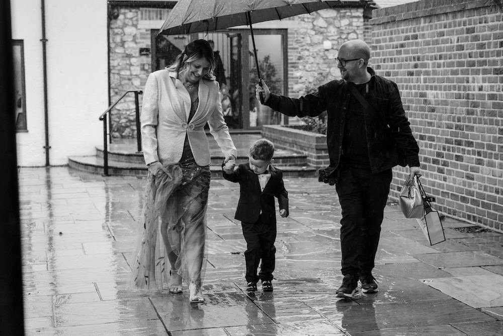 A woman and man are walking with a young boy on a rainy day, holding hands. The woman is wearing a light-colored blazer and dress, smiling, while the man holds an umbrella over them. The man has a camera and shopping bags, and they are walking on a wet sidewalk next to a brick wall.