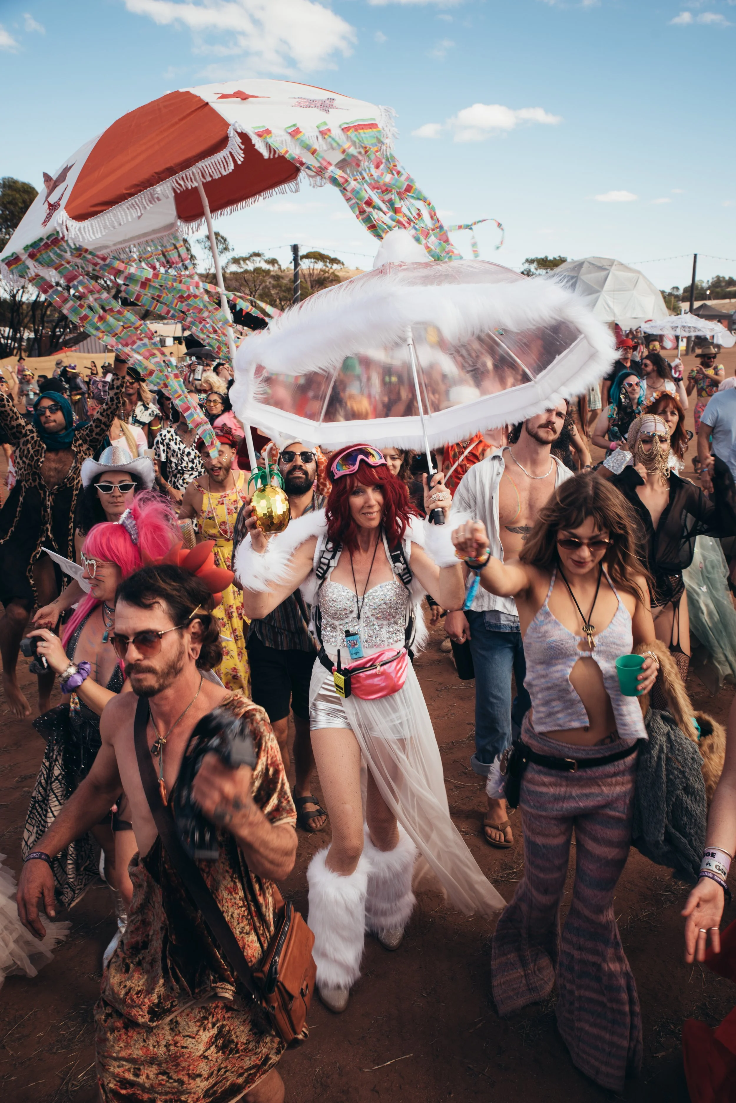 Crowd of festival attendees dressed in colorful and eccentric outfits, holding umbrellas, and walking outdoors on a sunny day.