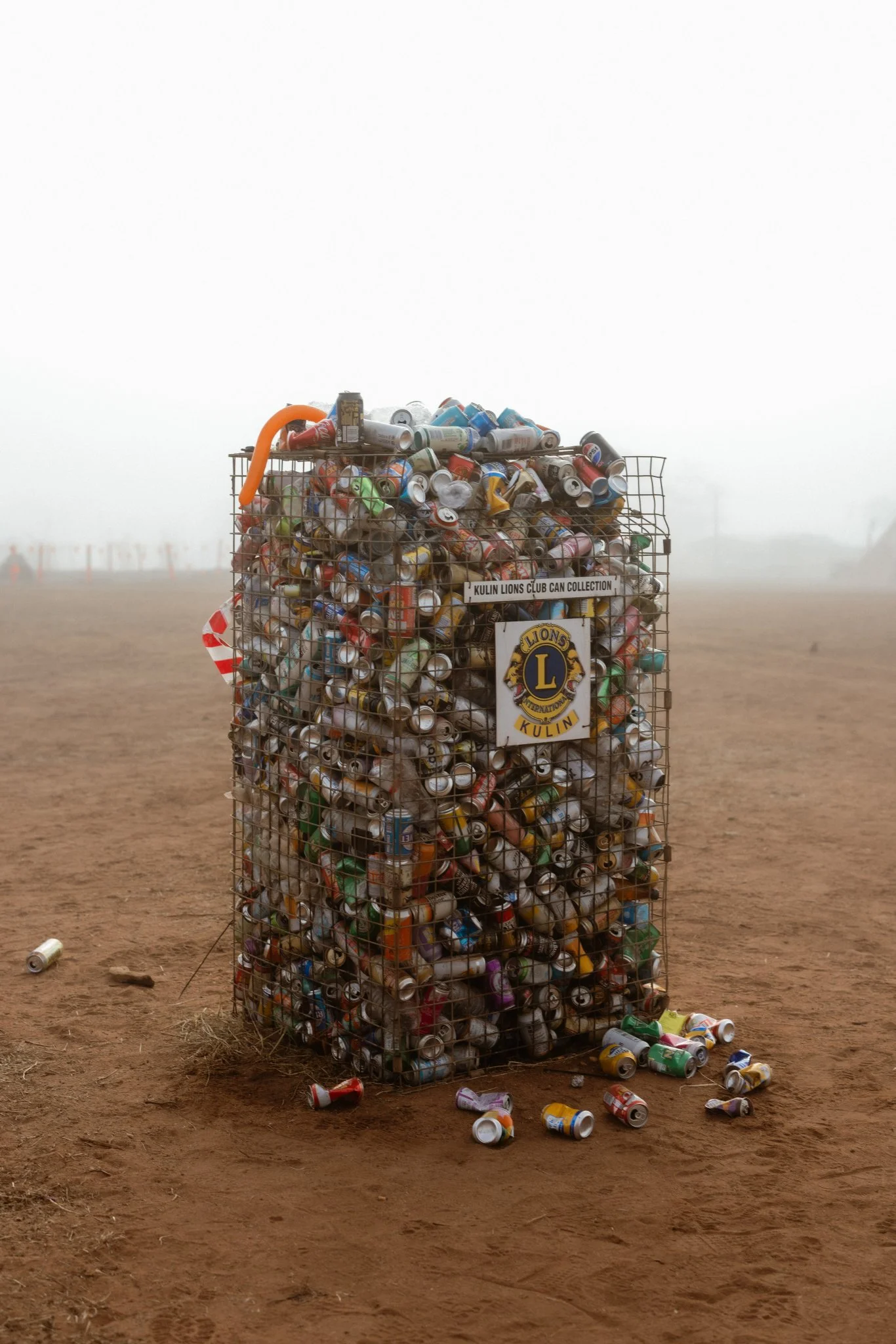 A wire cage filled with crushed cans, some scattered on the ground, labeled 'Kulin Lions Club Can Collection' on a dusty outdoor field with foggy weather.