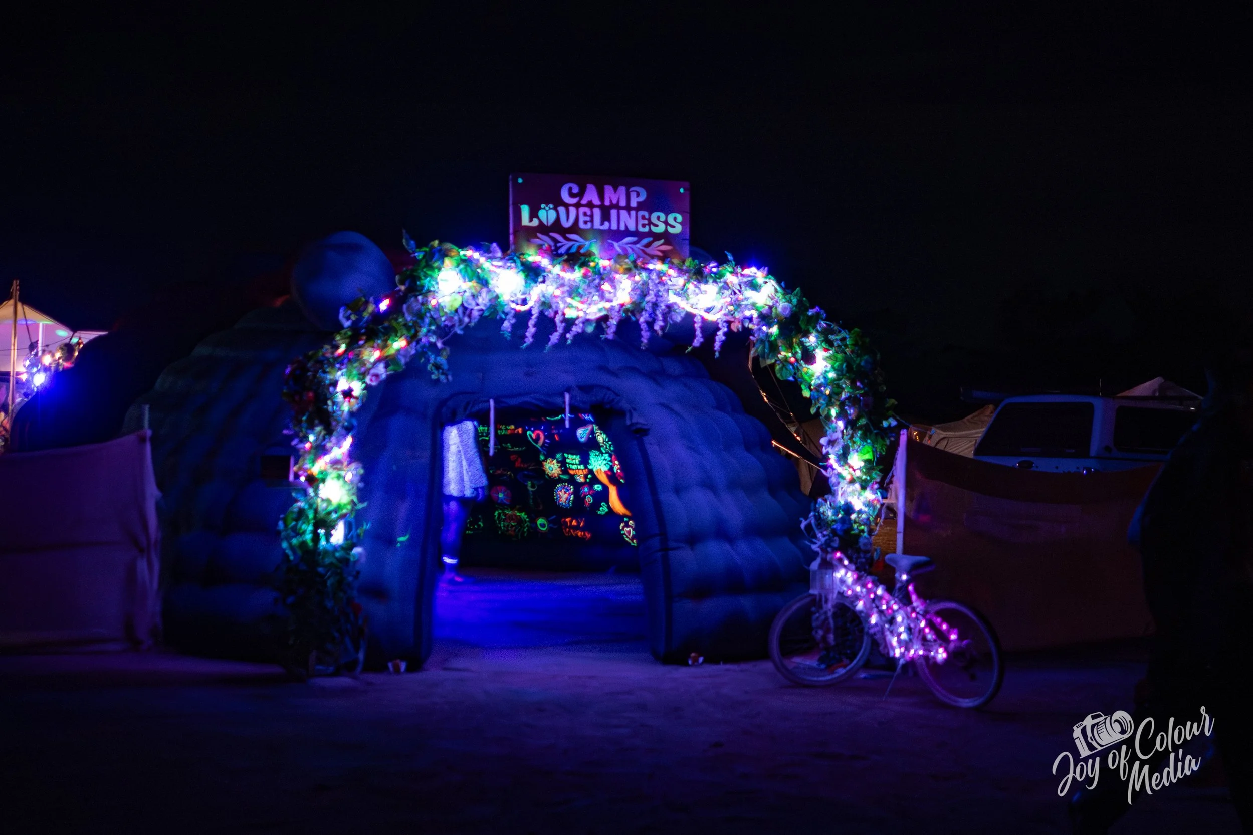 Night scene at Camp Loveliness with a decorated entrance arch illuminated by colorful lights. A sign reading 'CAMP LOVELINESS' is visible at the top of the arch, and tents can be seen in the background.