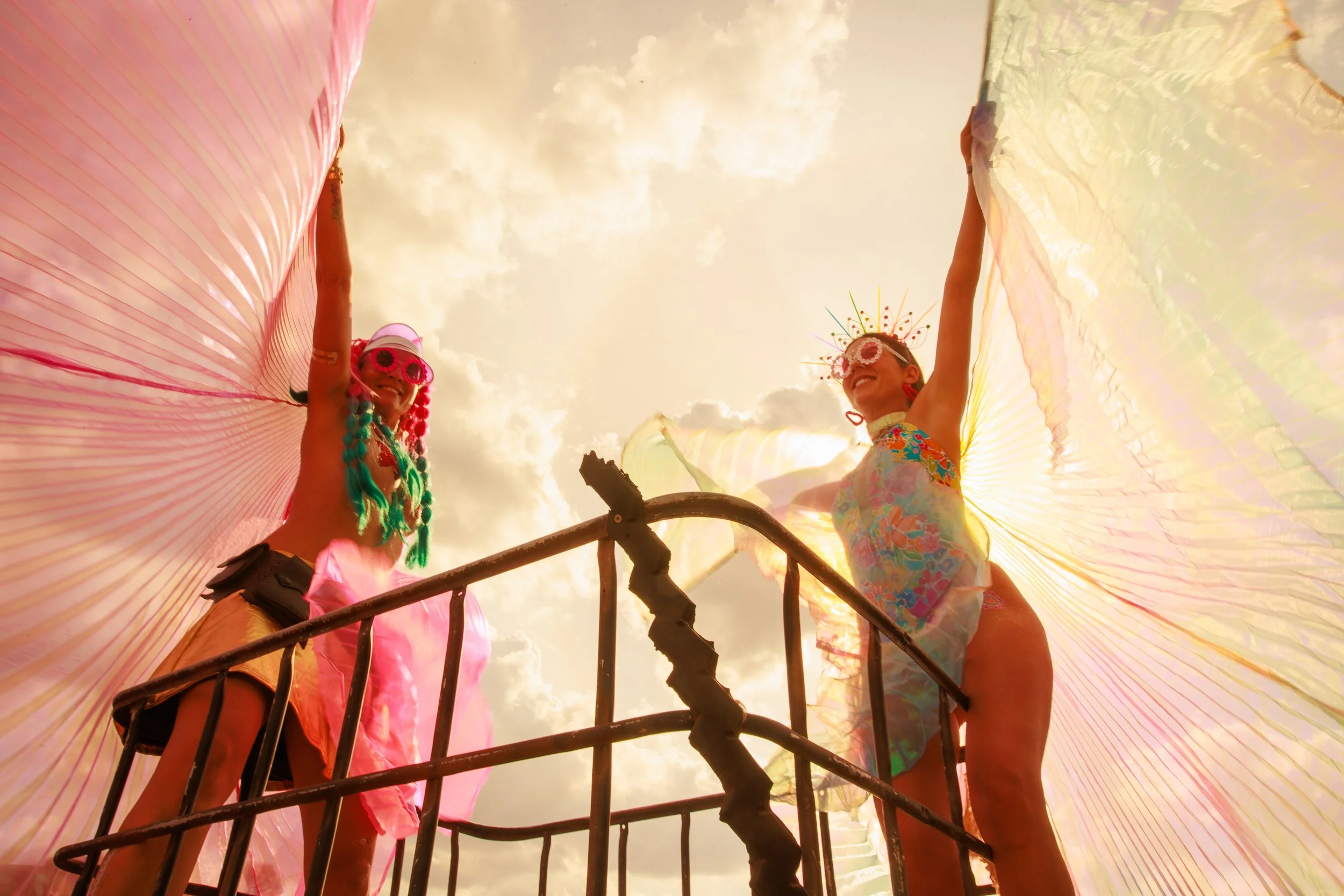 Two women dressed in colorful festival attire, wearing decorative glasses and headpieces, holding large colorful fans, standing on a metal platform outdoors with a cloudy sky in the background.