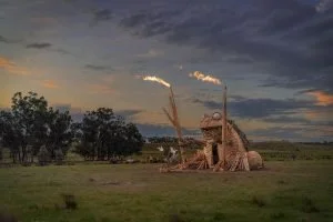 A large giraffe sculpture with smoke coming from its horns in a rural field at sunset.