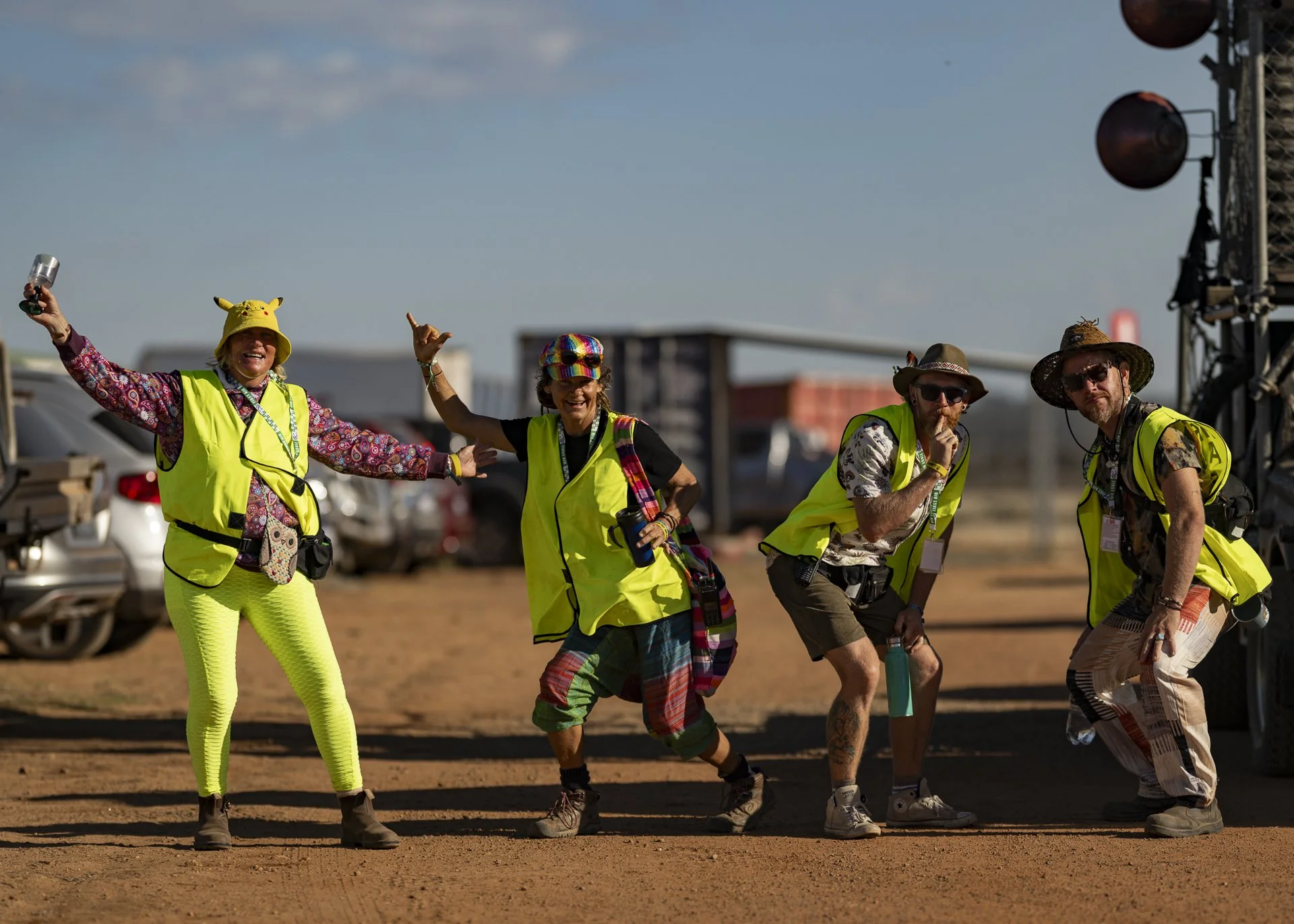 Group of people at an outdoor event, some wearing bright yellow safety vests, colorful hats, and casual clothing, standing on a dirt ground with cars and a stage with sound equipment in the background.