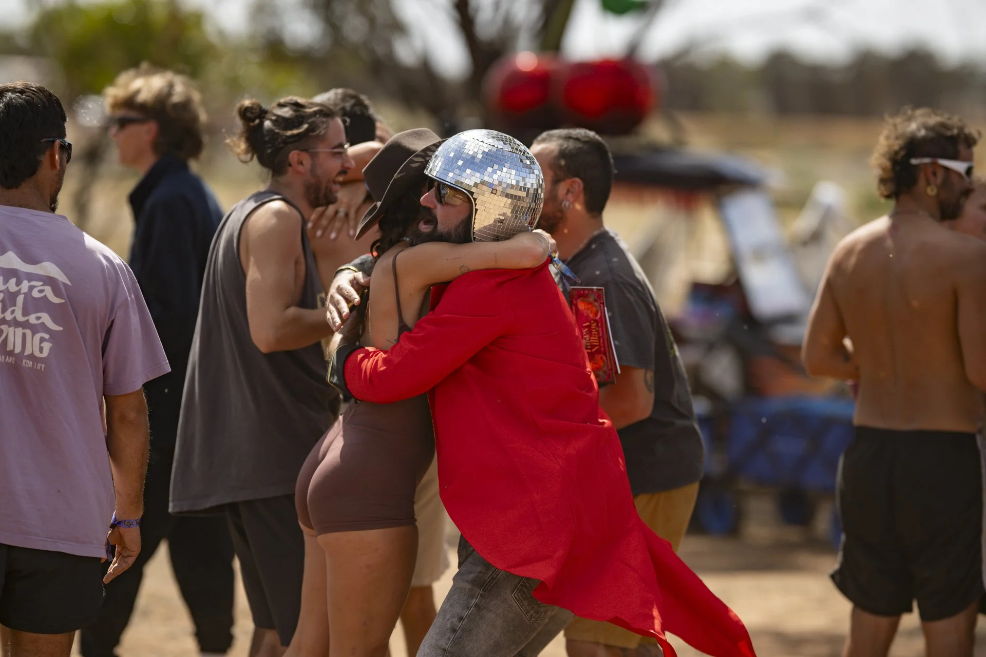 People hugging at an outdoor event, some wearing sunglasses and a shiny disco ball hat, with trees and vehicles in the background.