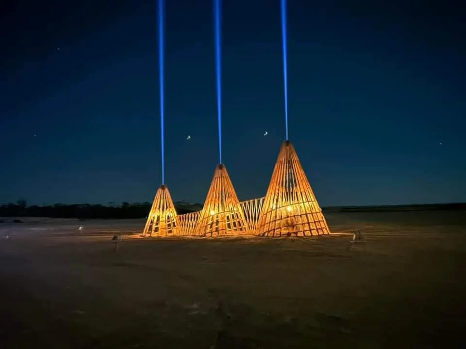 Nighttime view of illuminated sculptural installations resembling wooden cone-shaped structures with blue laser beams projecting upwards from the top.
