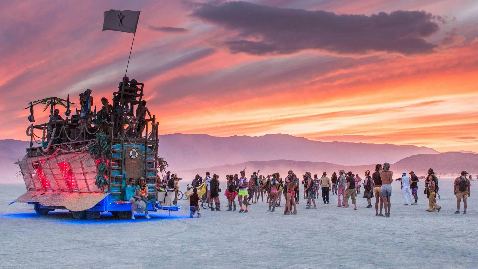 A large crowd of people gathered at sunset around a large wooden art installation on wheels, resembling a pirate ship with statues and a flag, in a vast open desert with mountain ranges in the background.