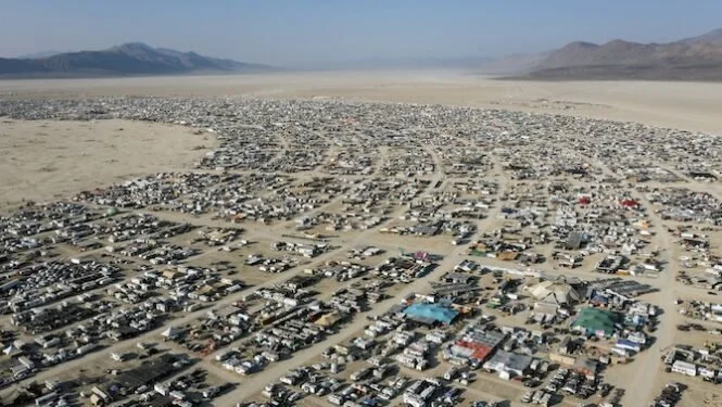 Aerial view of a large desert city with numerous vehicles and buildings, surrounded by mountains in the background.