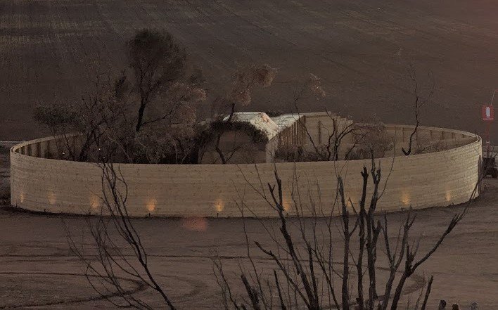 A circular wooden fence surrounds a small white building and some leafless trees, set against a dark, dirt landscape at night.