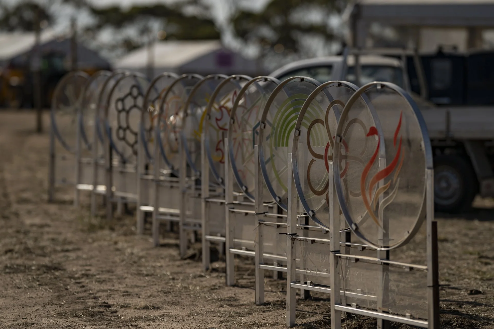 A row of bike racks with some painted designs on the side, outdoors in a dirt area, with a vehicle and trees in the background.