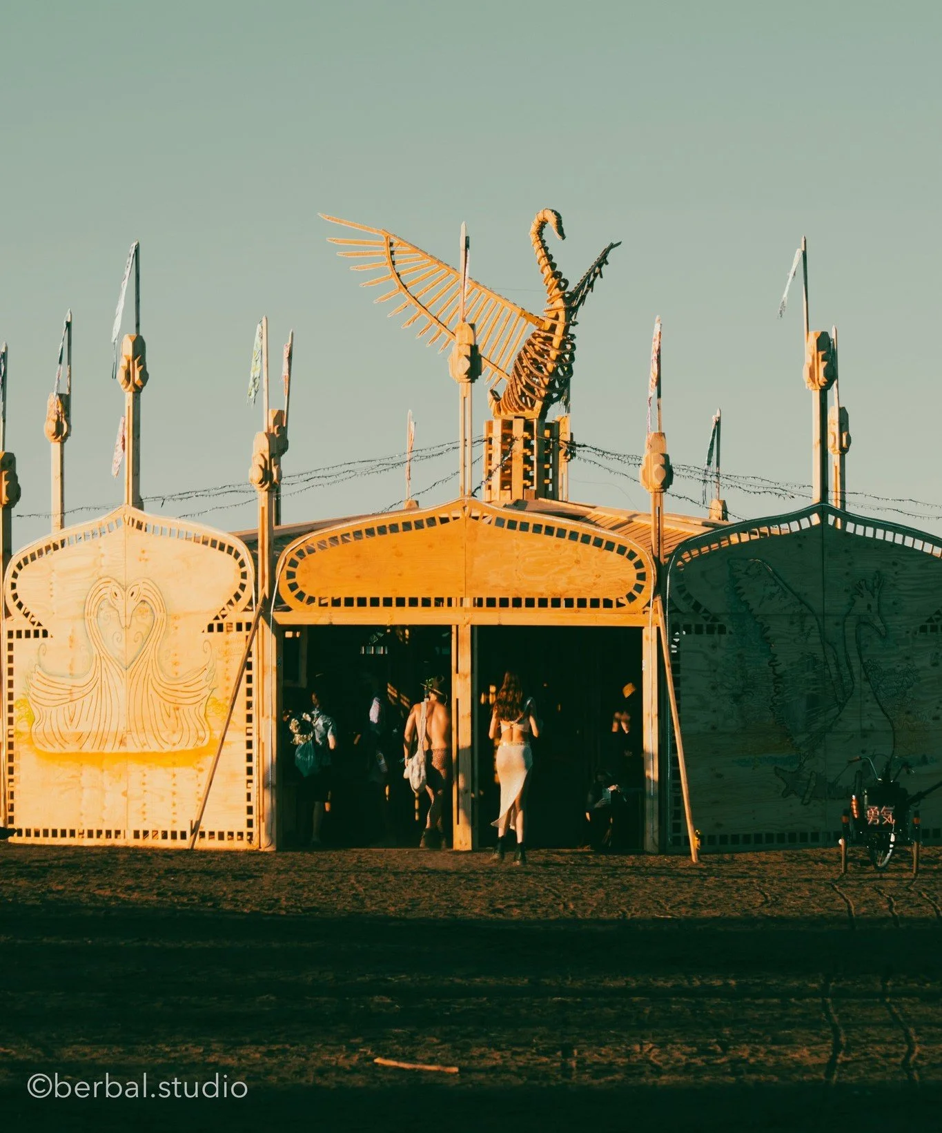 Wooden fairground ride with dragon and peacock decorations, people entering and exiting, under a clear sky.