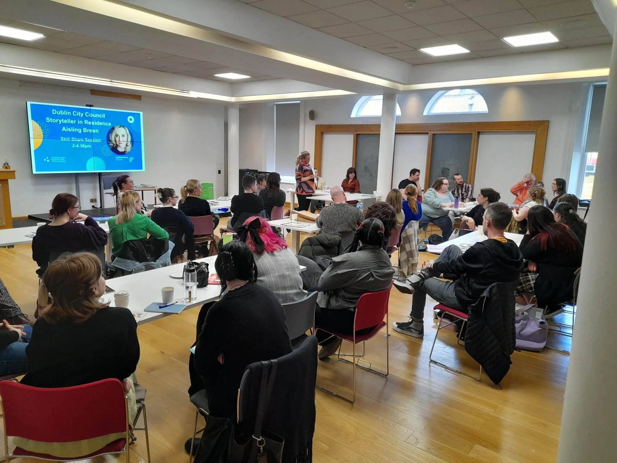 A presentation or meeting in a conference room with attendees seated around tables, a speaker at the front, a large screen displaying information about a storyteller in residence, Aisling Breen, and some windows with natural light.