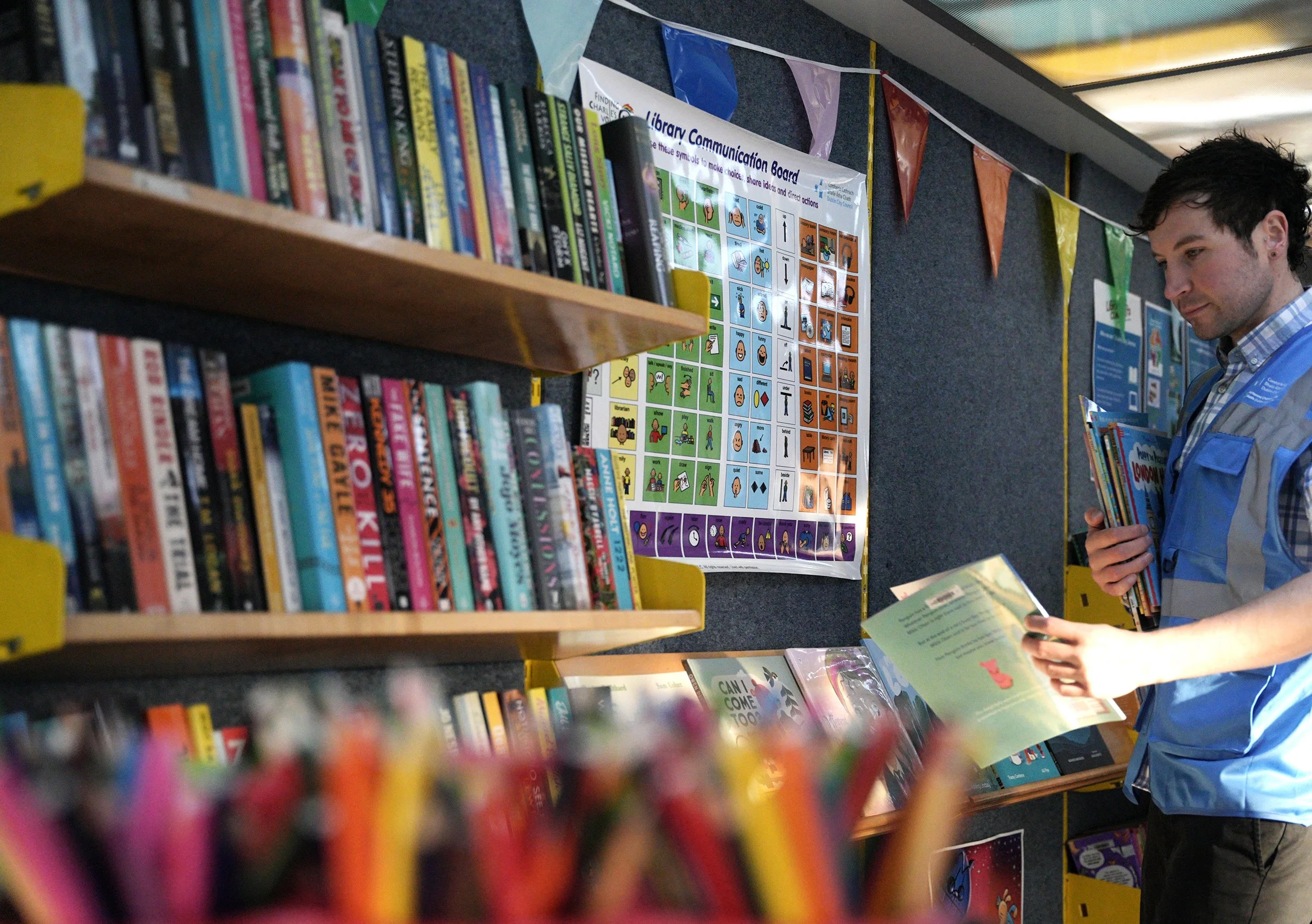 A person in a blue and gray vest is looking at children's books in a library, with shelves of books and a colorful communication poster in the background.