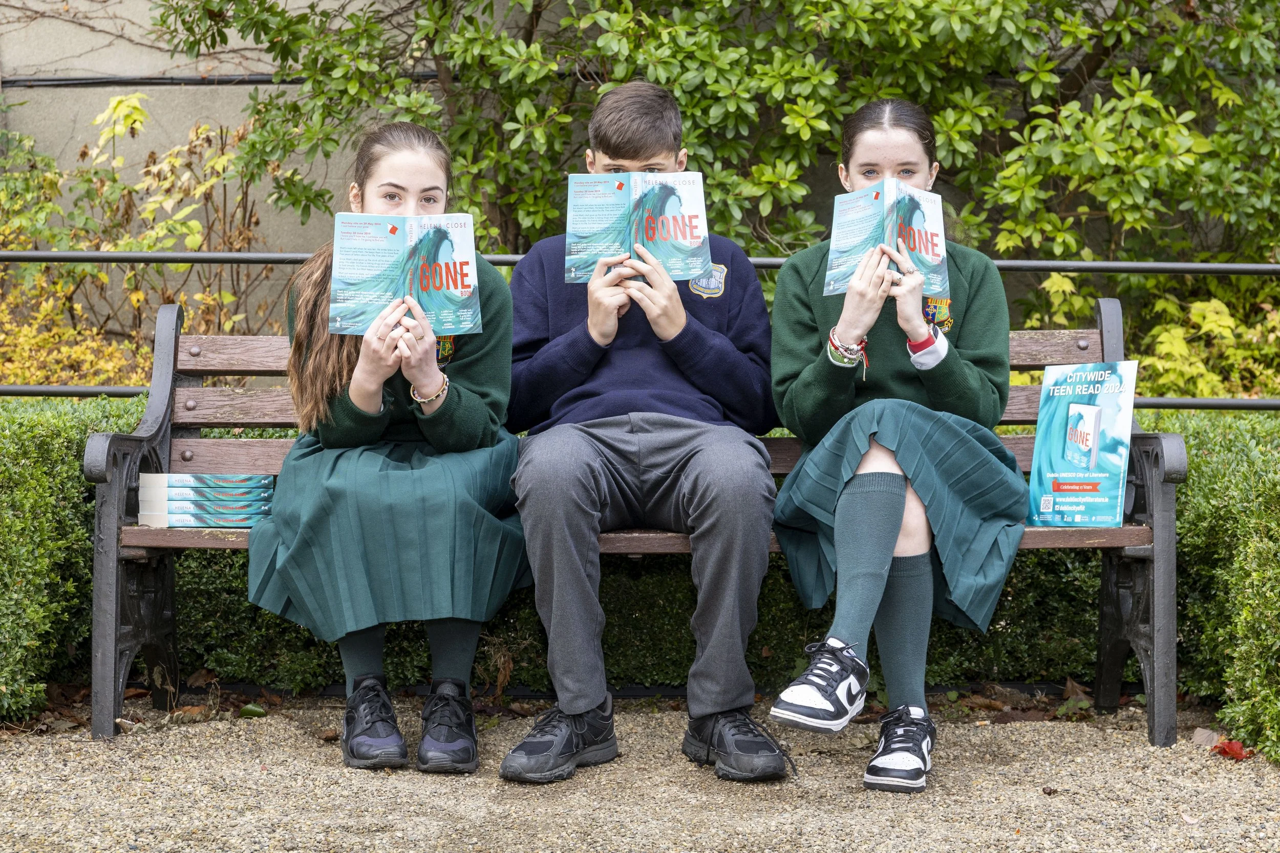 Three teenagers sitting on a park bench, holding books in front of their faces, with a background of greenery. The books feature a blue cover with the word 'GONE' in red. A sign on the bench reads 'Citywide Teen Read 2024.'