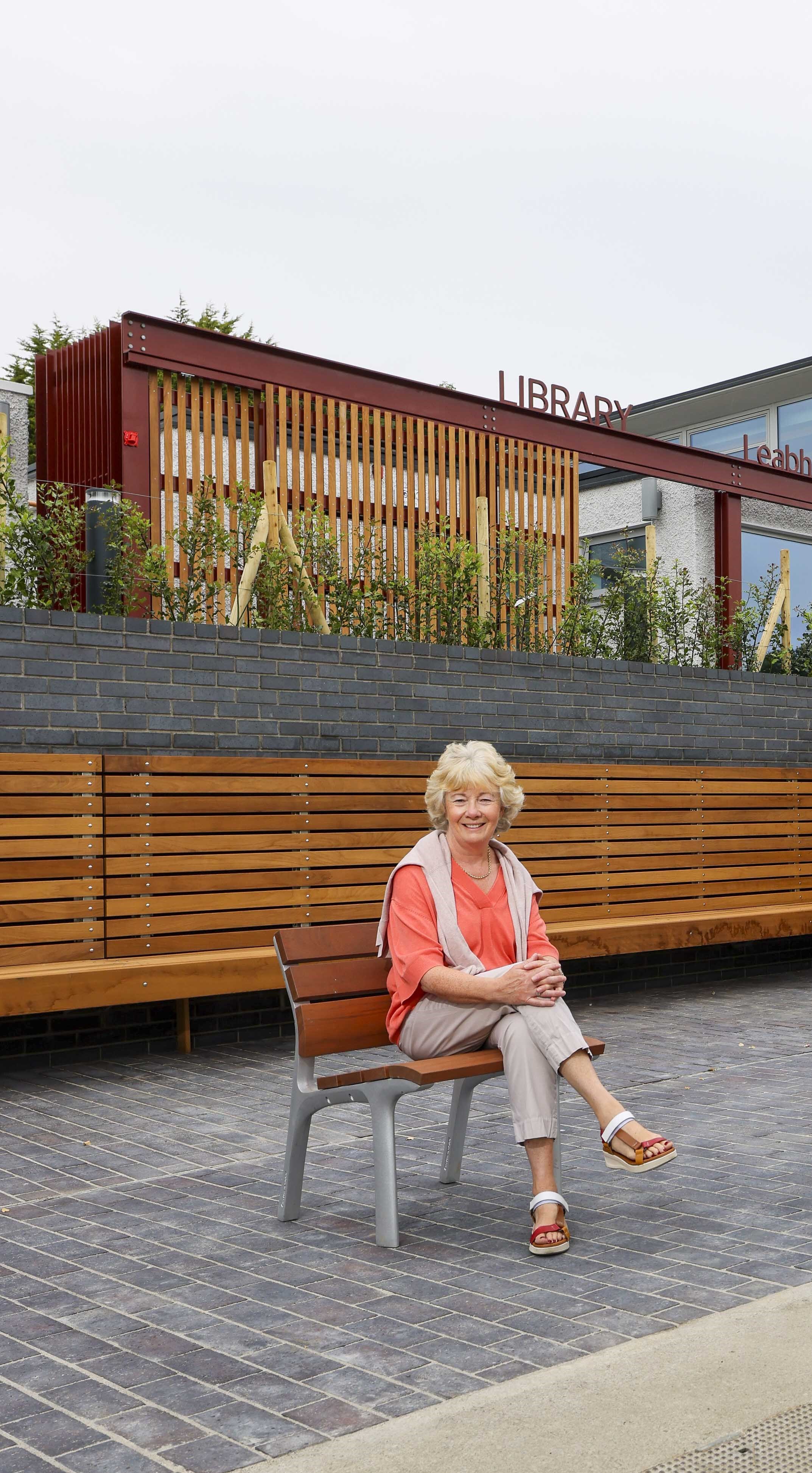 An older woman sitting on a wooden bench in front of a modern building with a sign that reads 'Library'. She is smiling, wearing a coral top, beige pants, and sandals.
